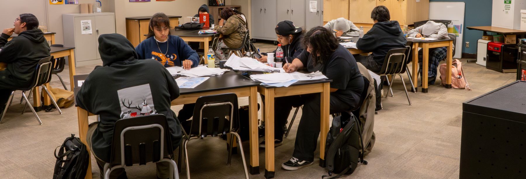 Students in a school classroom focused on their homework at desks, with books and supplies scattered around them.